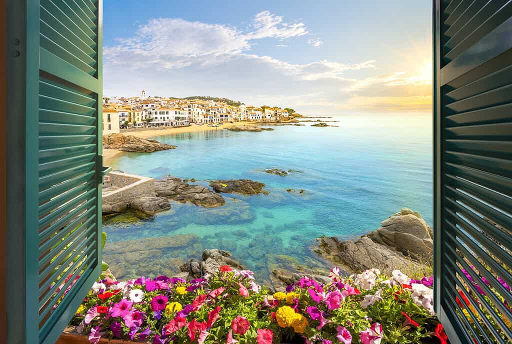 View through an open window with shutters of the sandy beach, rocky coastline and whitewashed town of Calella de Palafrugell, Spain, on the Costa Brava coast as the sun sets. The best summer activities for kids in the Costa Brava.